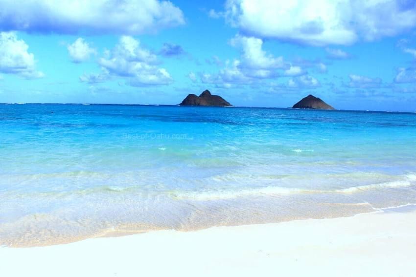 A serene beach scene with clear blue water, white sand, and distant islands under a partly cloudy sky.