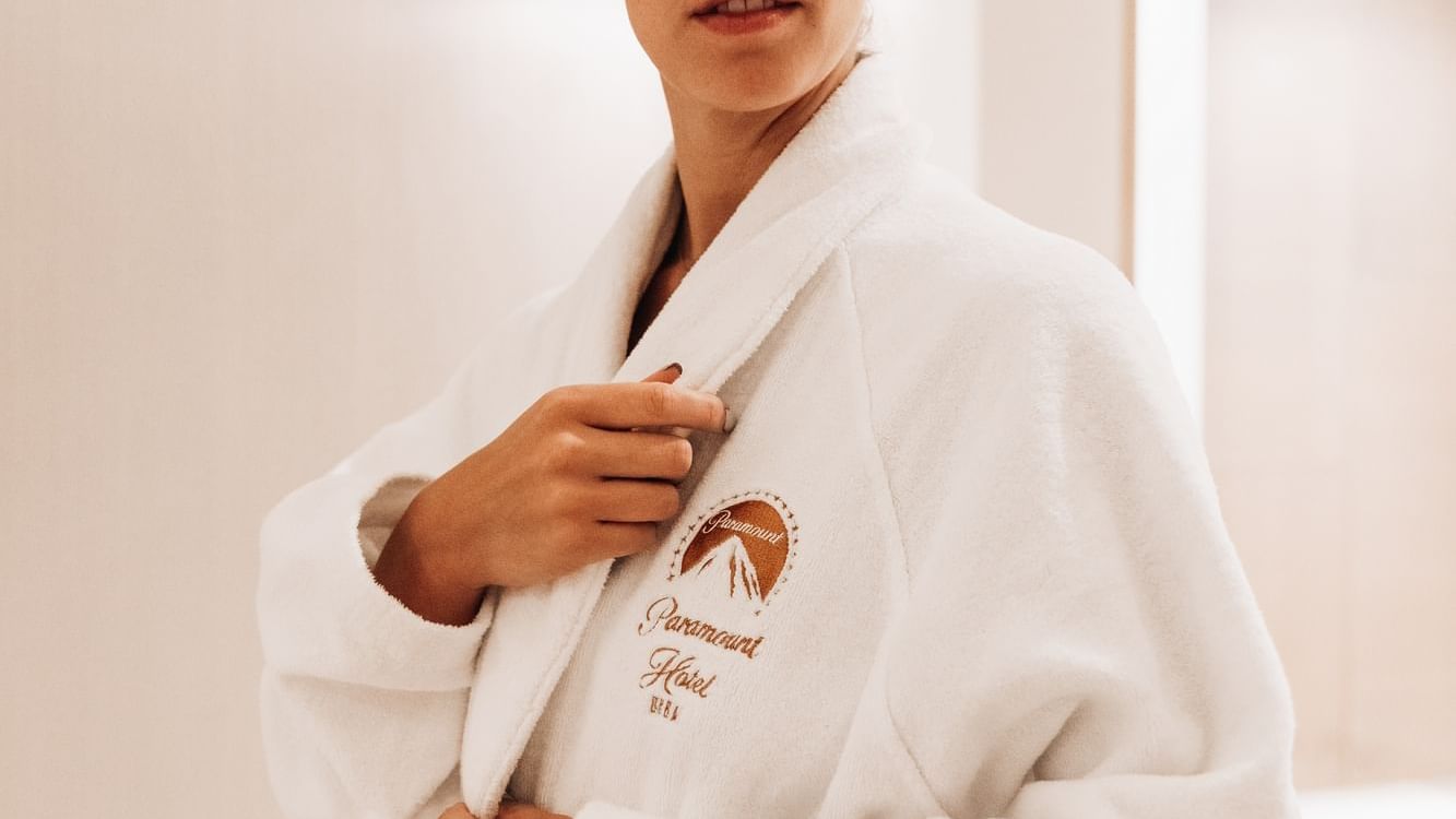 Lady in a plush white Paramount Hotel bathrobe stands in a softly lit, modern interior at Paramount Hotels