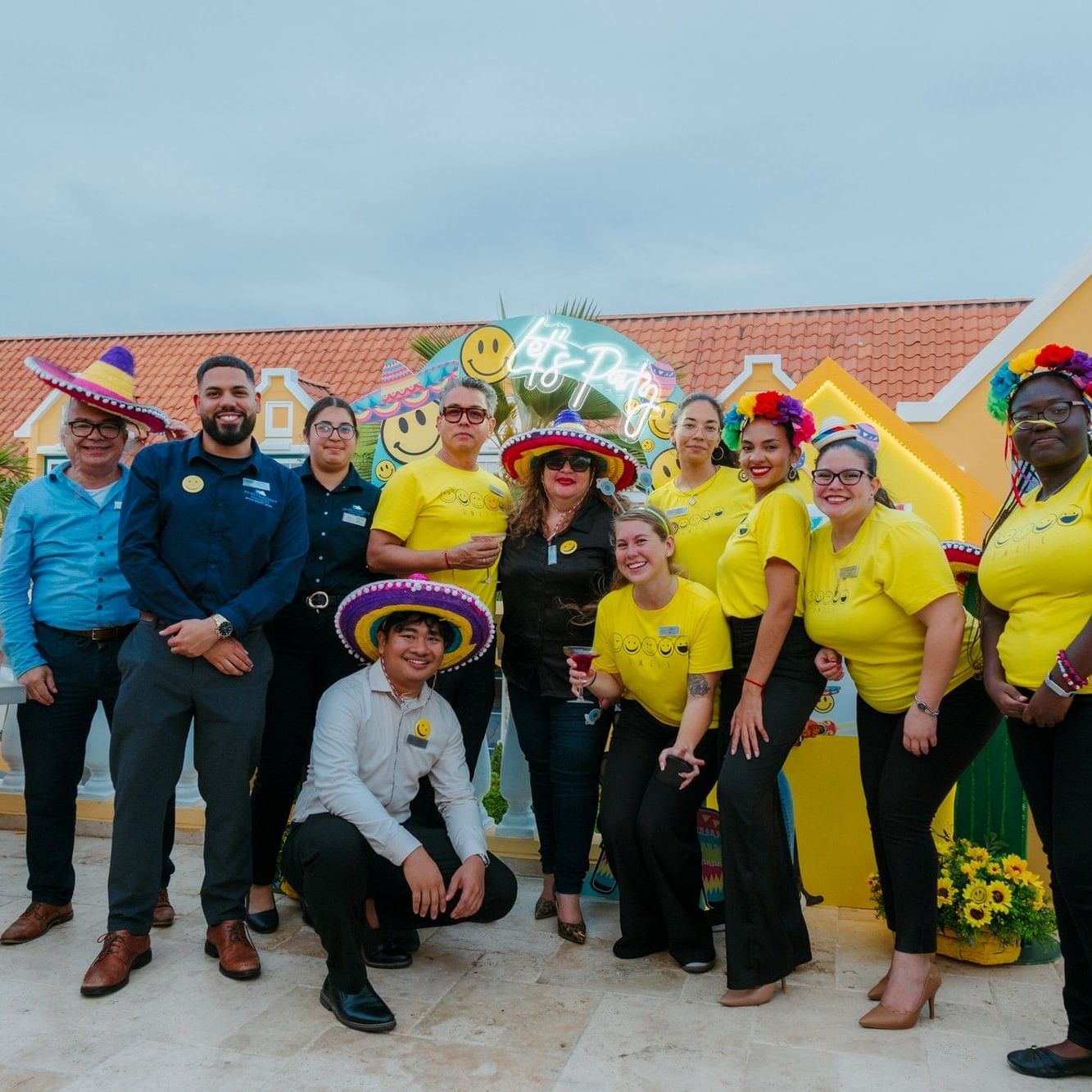 Resort staff celebrating with colorful smiley face balloons and festive sombreros on a sunny terrace in Aruba.