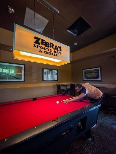 A man shoots pool beneath a hanging sign that reads Zebra's Sports Bar & Grill. Zebra's is a great place for Karaoke in Orlando.