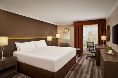 Guestroom with white bed, nightstands, desk, chair, TV, and window with curtains showing an outdoor view.