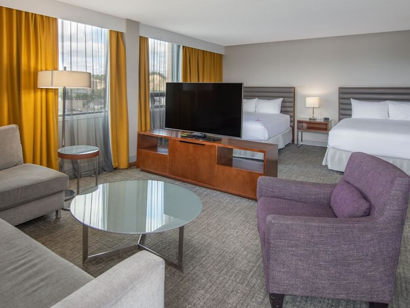 A hotel suite with a couch, chair, coffee table, TV, and two queen beds.