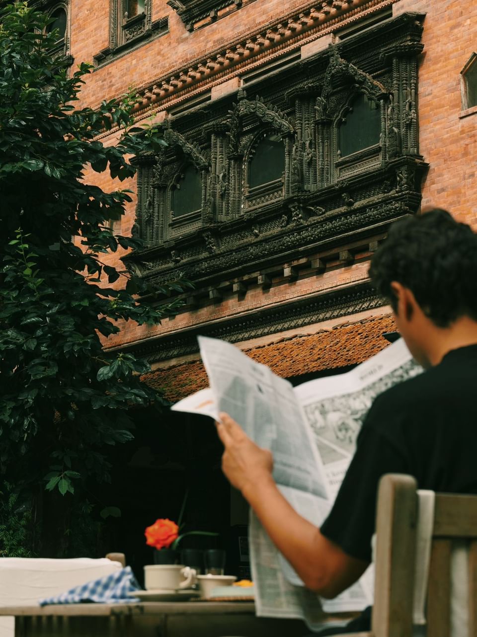 A person sits at a table with a newspaper, coffee, and a flower at The Dwarika’s Hotels and Resorts