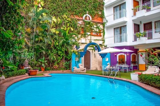 Colorful courtyard pool beside patio table & chairs surrounded by lush greenery with a chapel facade at Los Arcos Suites