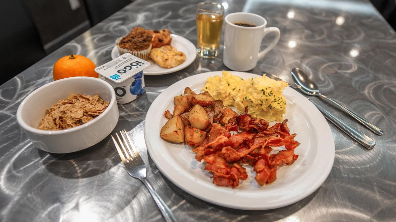 Breakfast spread with scrambled eggs, bacon, potatoes, cereal, pastries, juice, and coffee on a patterned tablecloth