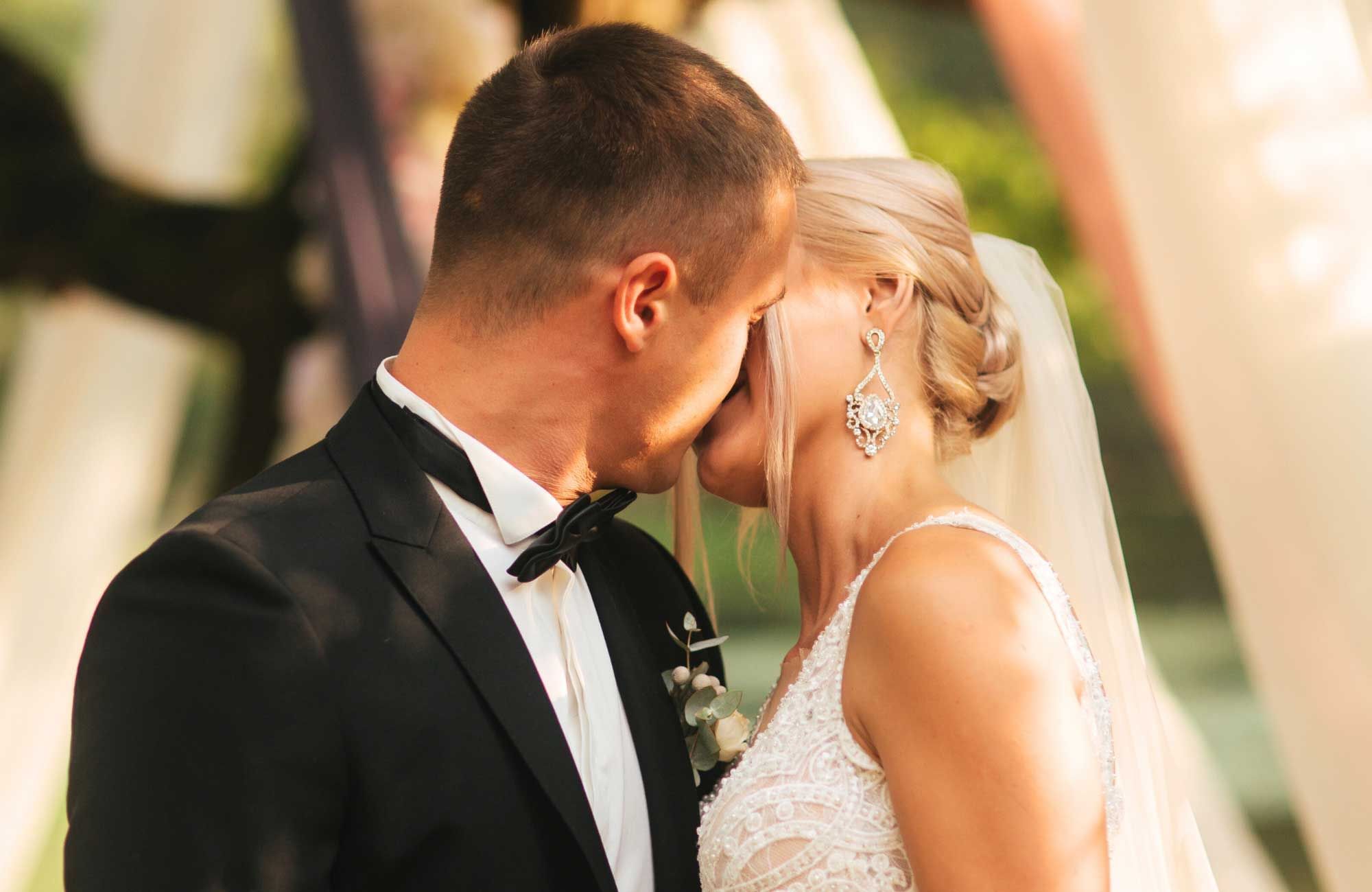 Bride in an elegant gown and earrings leans in for a kiss with her groom, surrounded by outdoor decor at Cove Pocono Resorts