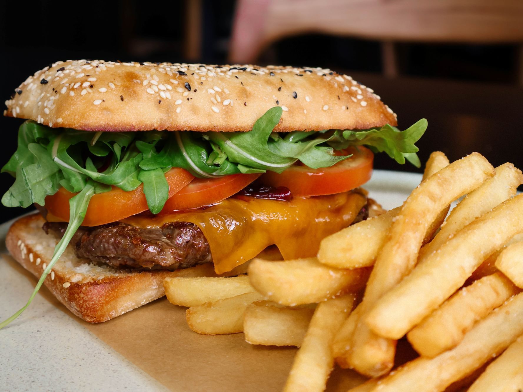 A gourmet steak sandwich with melted cheese, tomato, and rocket on a toasted bun, served with a side of golden fries on a white plate, as part of The Weekender package at Hotel Grand Chancellor Townsville.