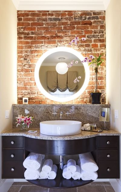 Bathroom vanity with a lit circular mirror against brick wall in Inn at Willow Grove