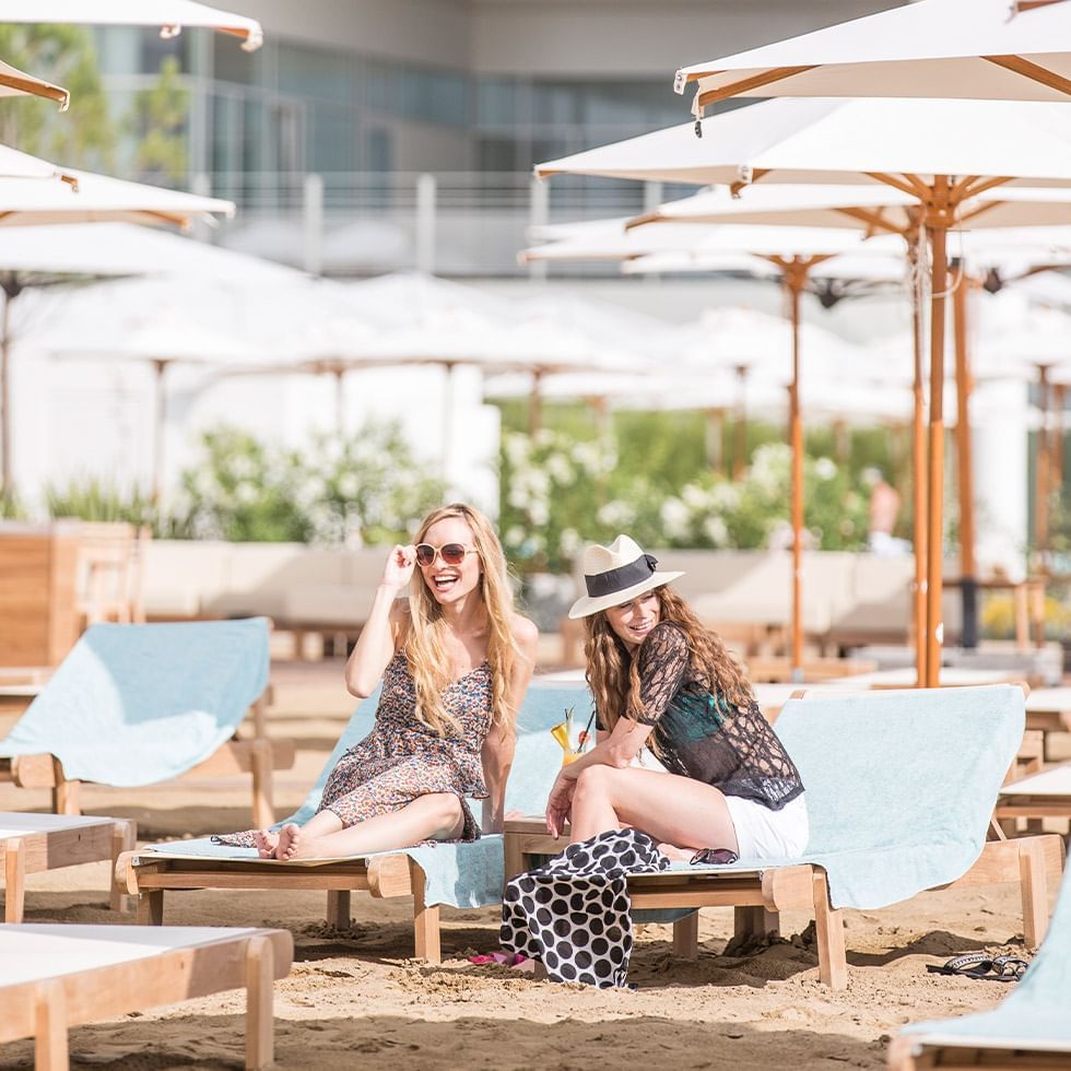 Zwei Frauen in Strandkleidung sitzen auf Liegen unter Sonnenschirmen neben einem Gebäude.