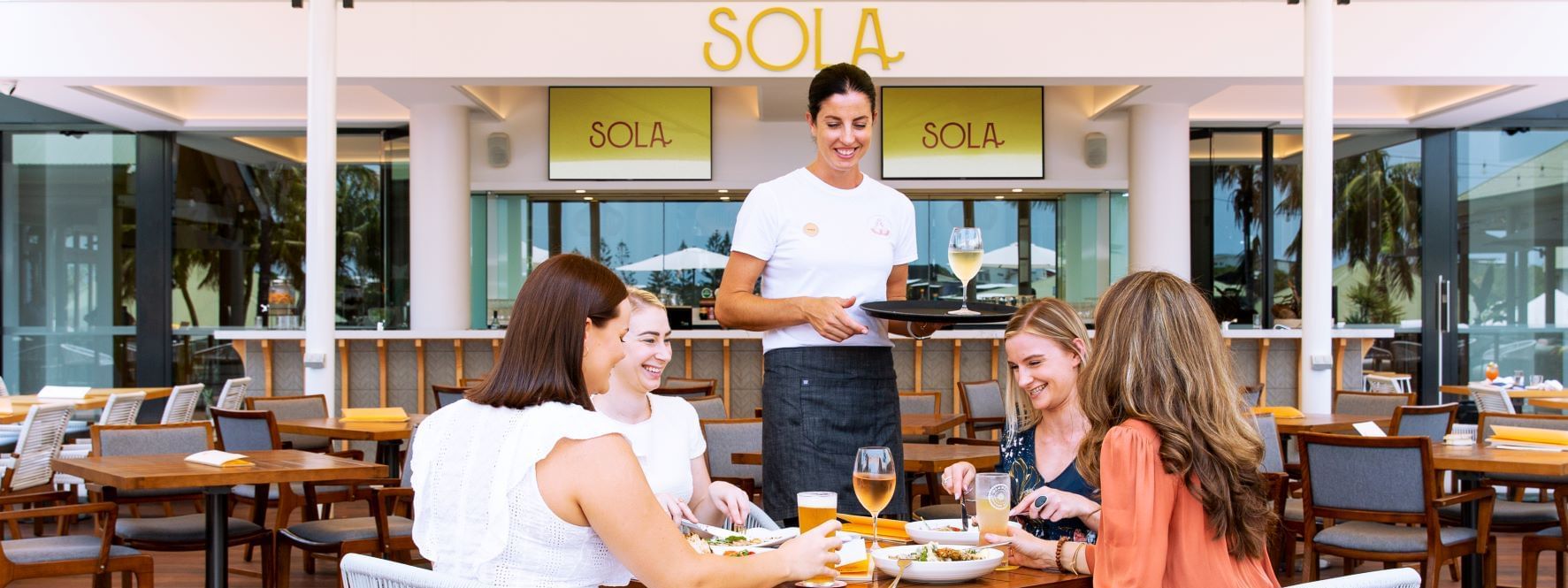 Waiter serving food to four women in Sola outdoor bar with modern decor and tropical plants at Novotel Sunshine Coast Resort