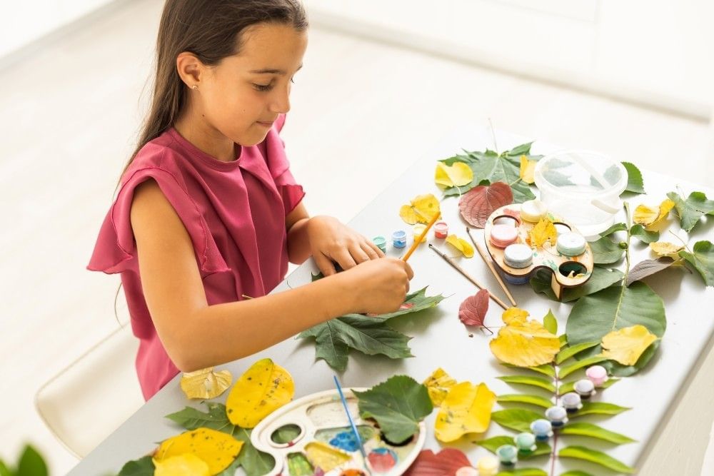 A girl sits at a table of leaves and colorful craft supplies. 
