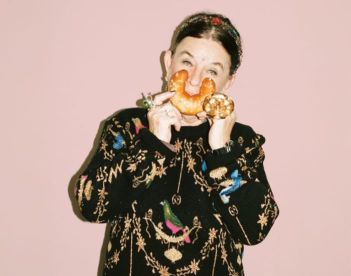 Marianne Kohn in a colorful black sweater holding a Kipferl (Austrian croissant) against a pink wall at Hotel Motto Vienna