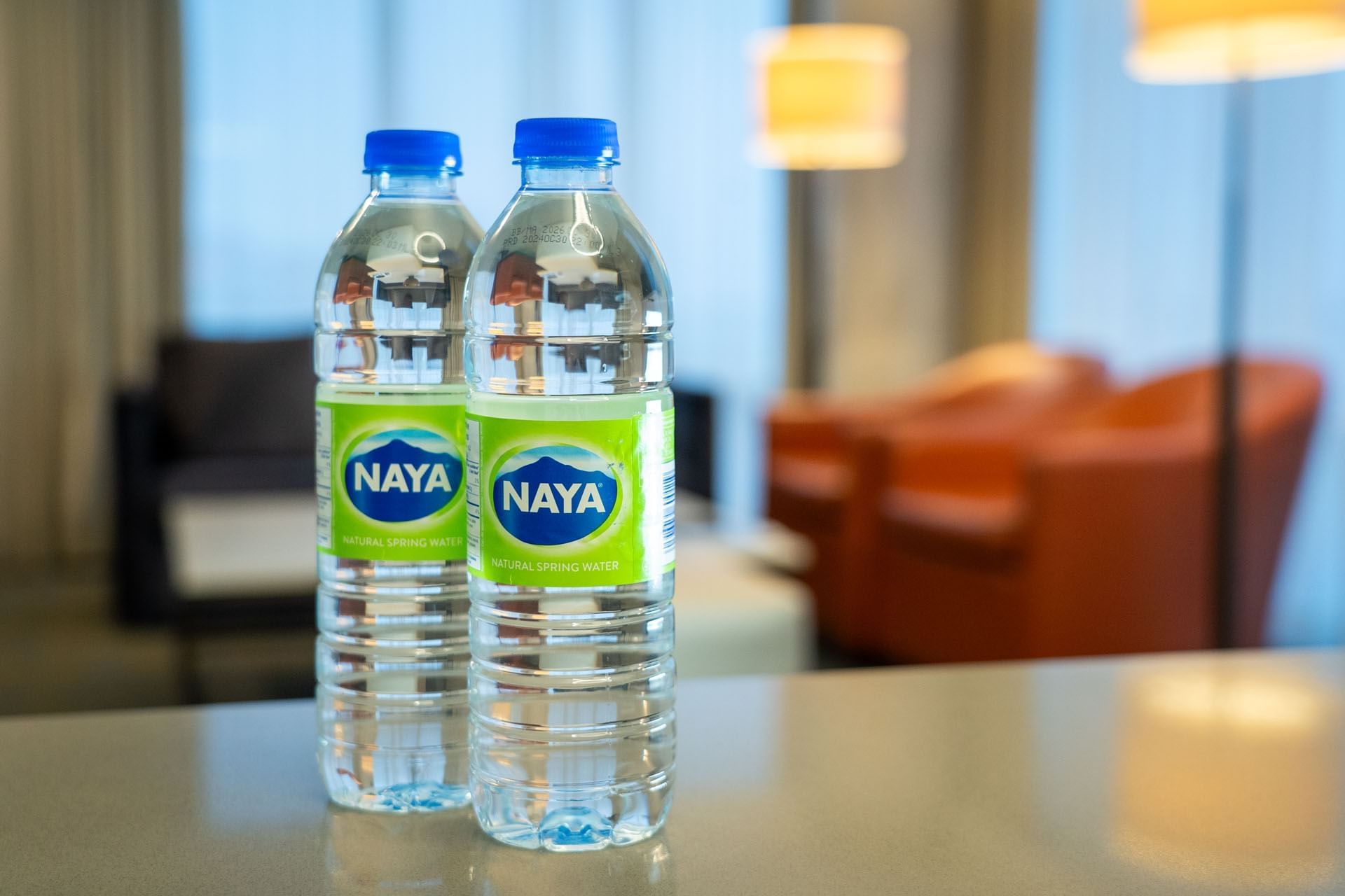 Two NAYA water bottles on a counter by orange armchairs and a glowing floor lamp in a blurred lounge at Hotel X Toronto