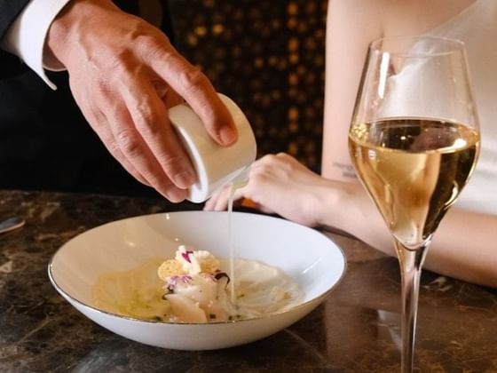 Hand pouring sauce over an elegantly plated dish, while a woman’s hand rests nearby in Square One at Park Hyatt Saigon