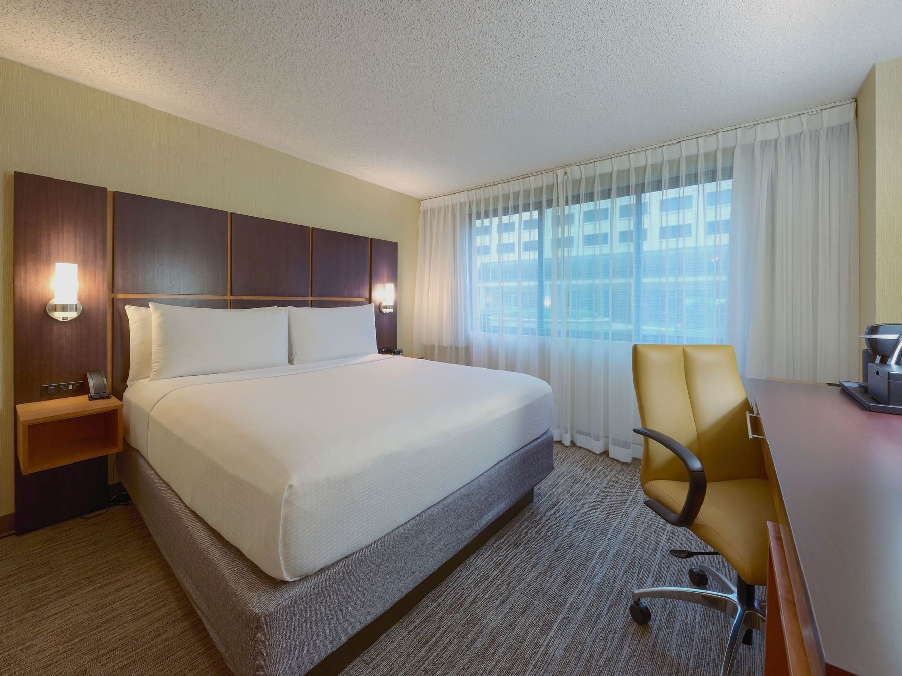 Classic King Room of the NCED Hotel and Conference Center with a bed, desk, chair, and window.