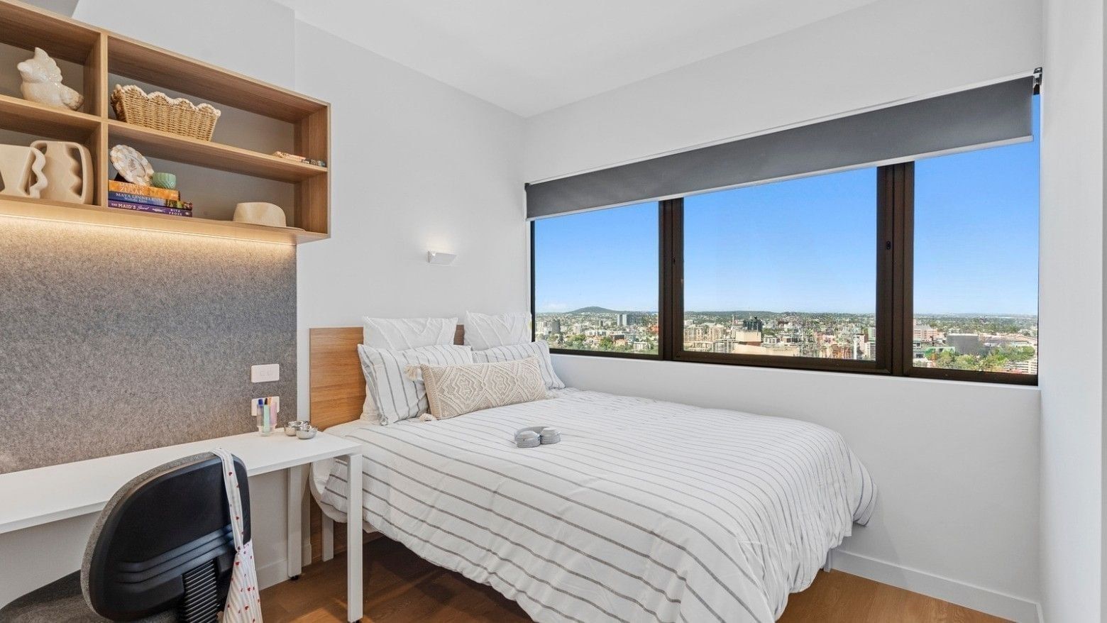Modern bedroom with white linens, wooden desk, open shelving, and large window showcasing city views at UniLodge Brisbane City