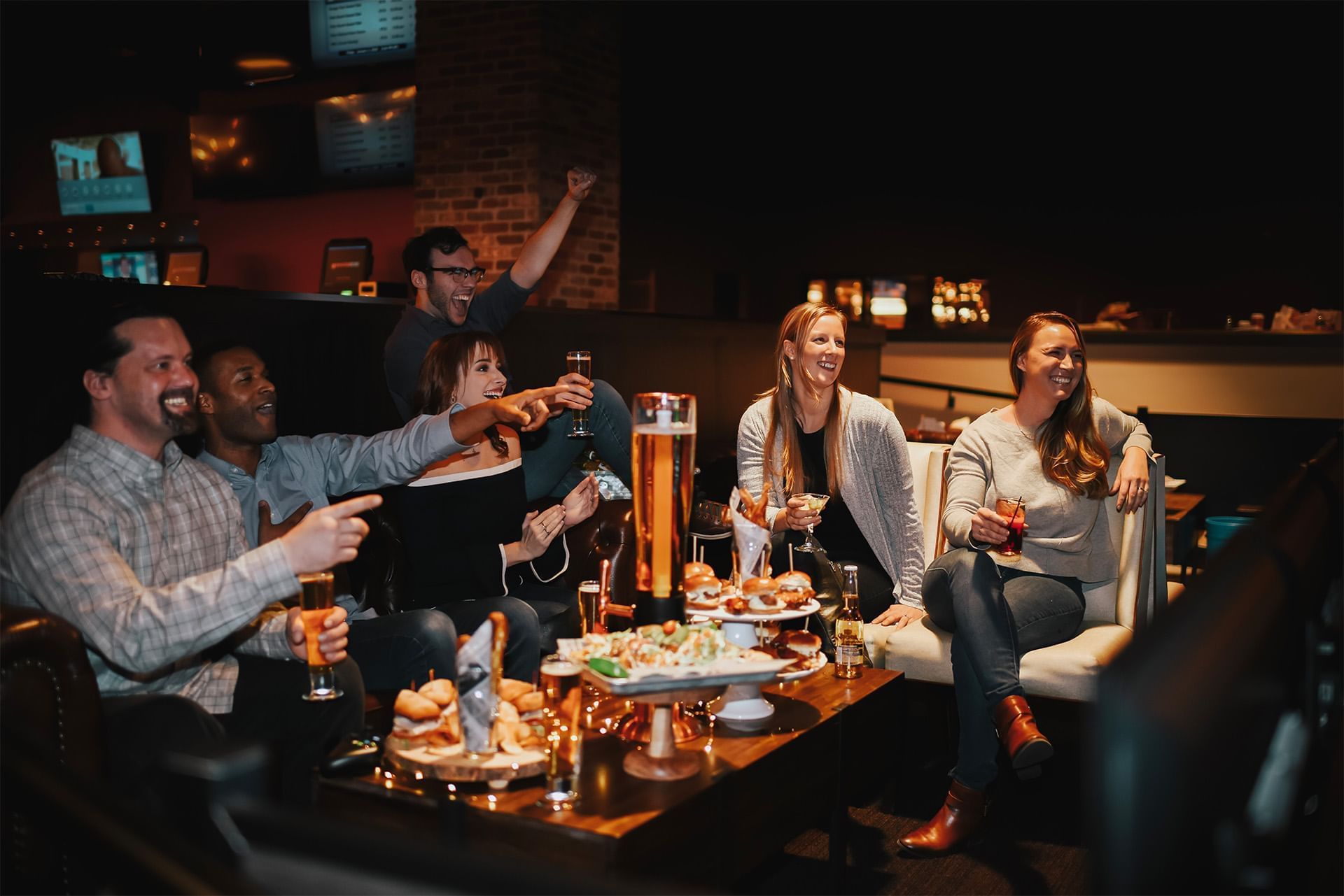 Friends cheer around a table with drinks and shared food while watching TV screens in the casino lounge at The Brook