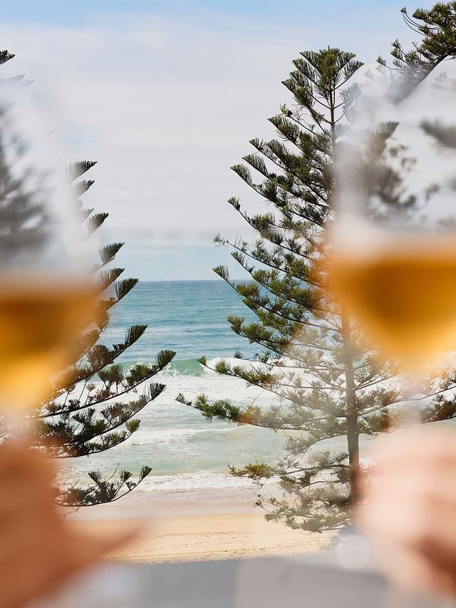 Pair of blurred wine glasses held by guests overlooking ocean waves and pine trees near the balcony at Elysium Manly