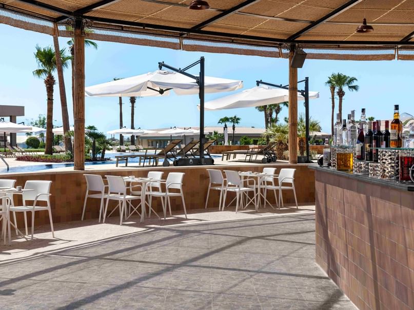 Outdoor patio with white tables, chairs, bar counter with bottles, and umbrellas near a pool and palm trees.