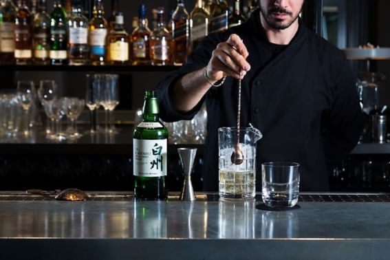 Bartender stirs a cocktail with ice in a glass while a bottle of Hakushu whiskey sits nearby on bar counter at Peter Street Kitchen