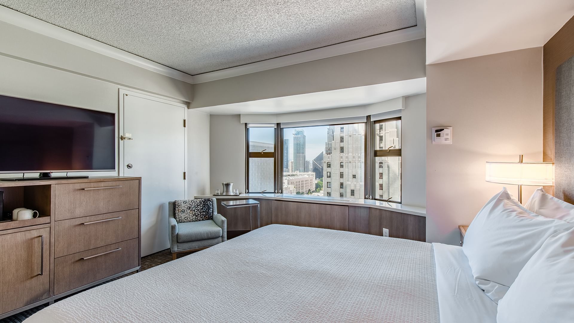 A hotel room with a king-size bed, TV, window with city view, chair, and lamp.