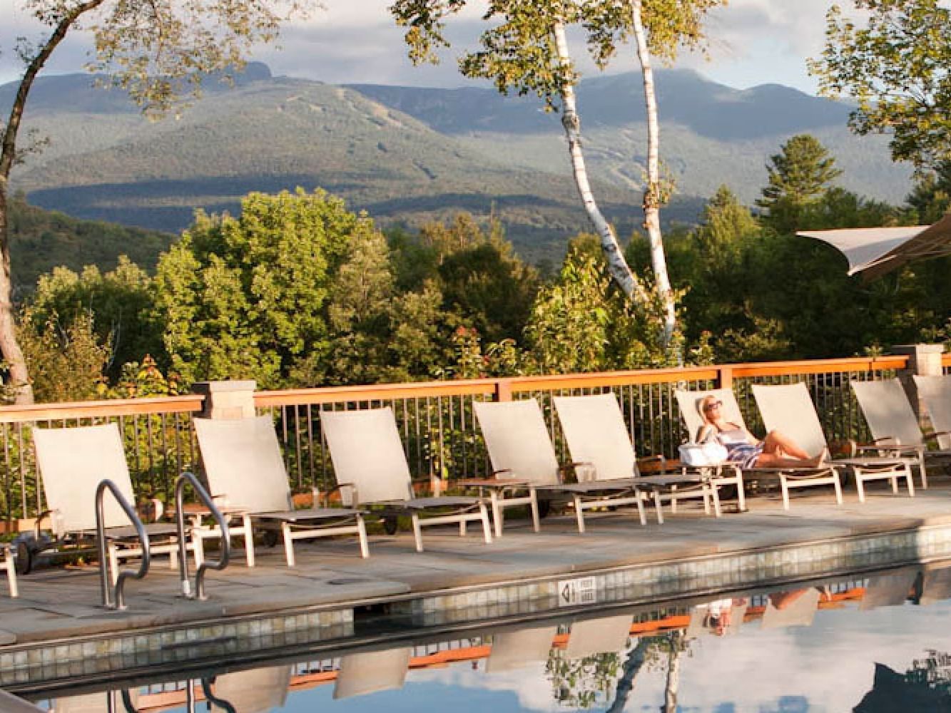 A person lounges on a chair by a pool with mountains in the background.