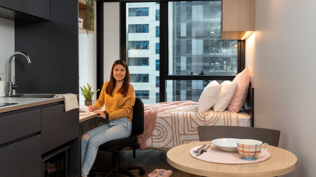 Resident sitting at desk in =studio apartment with kitchen, dining area and bed.