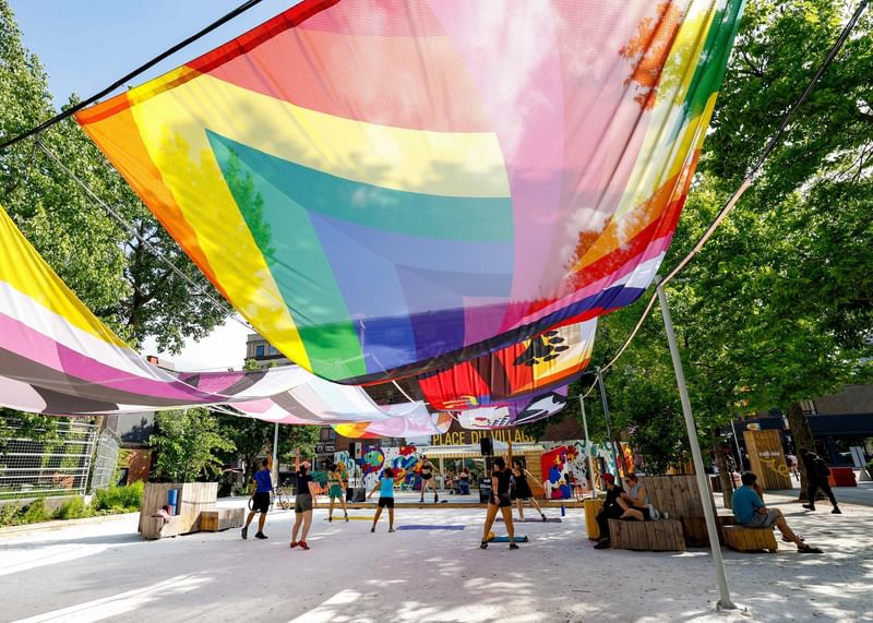 Rainbow pride flags hanging over an outdoor plaza with people relaxing on wooden benches at Warwick Le Crystal Montreal