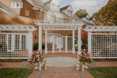 White pergola structure with floral arrangements and candles on stone patio beside brick exterior at The Simsbury Inn
