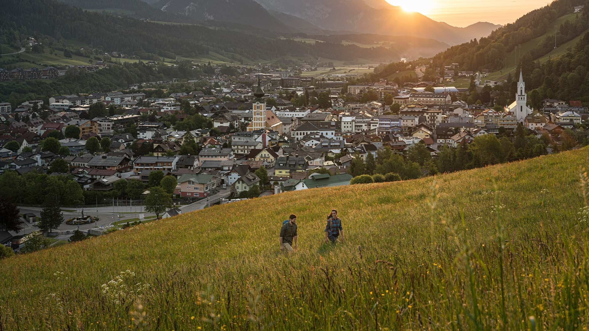 Zwei Wanderer auf einer Wiese über einer Stadt mit Alpenkulisse beim Sonnenuntergang.