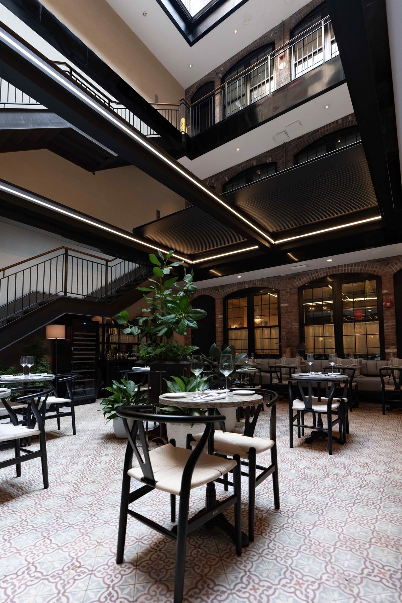 A dining area with tables and chairs, plants, a staircase, and windows.