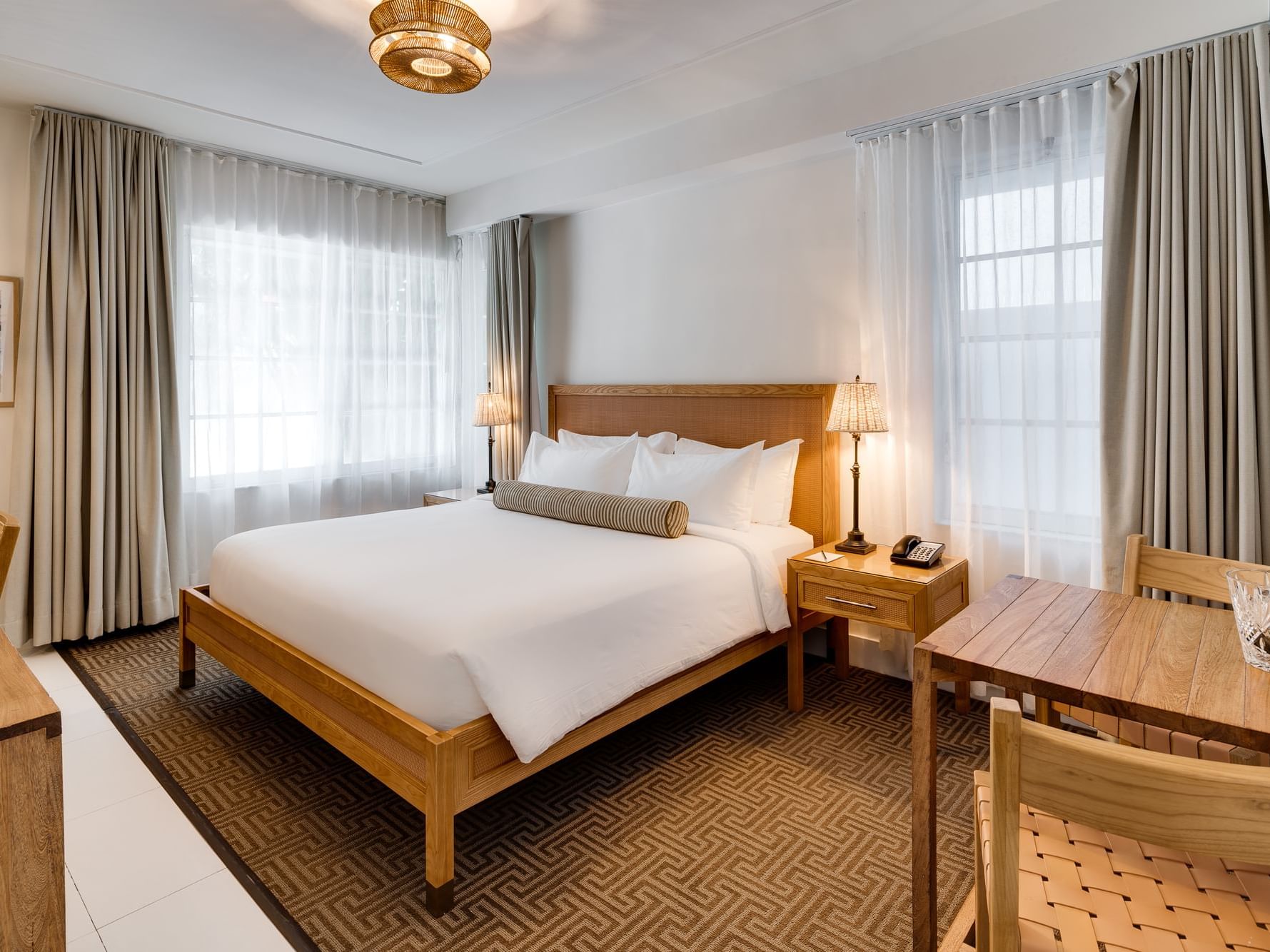 Newly renovated hotel room featuring a King bed with crisp white linens and a striped bolster pillow, surrounded by beach chic decor. The room includes wood furnishings, two bedside lamps, a writing desk, a dining table with chairs, and large windows with