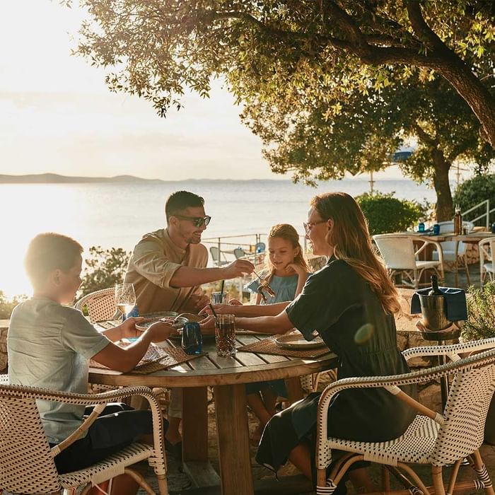 Famiglia seduta attorno a un tavolo da pranzo all'aperto con vista sul mare al tramonto.