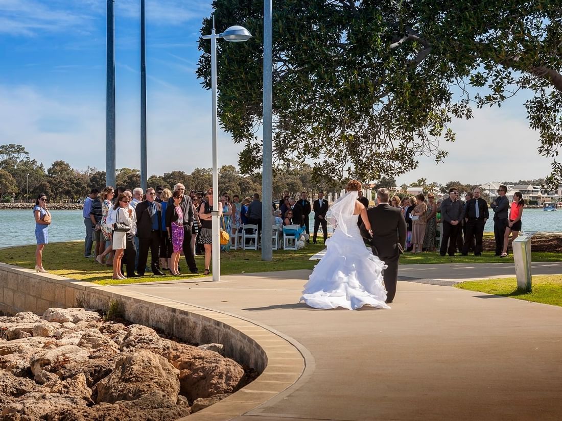 Bride and groom walk along an estuary path as wedding guests gather on a sunny day at The Sebel Mandurah