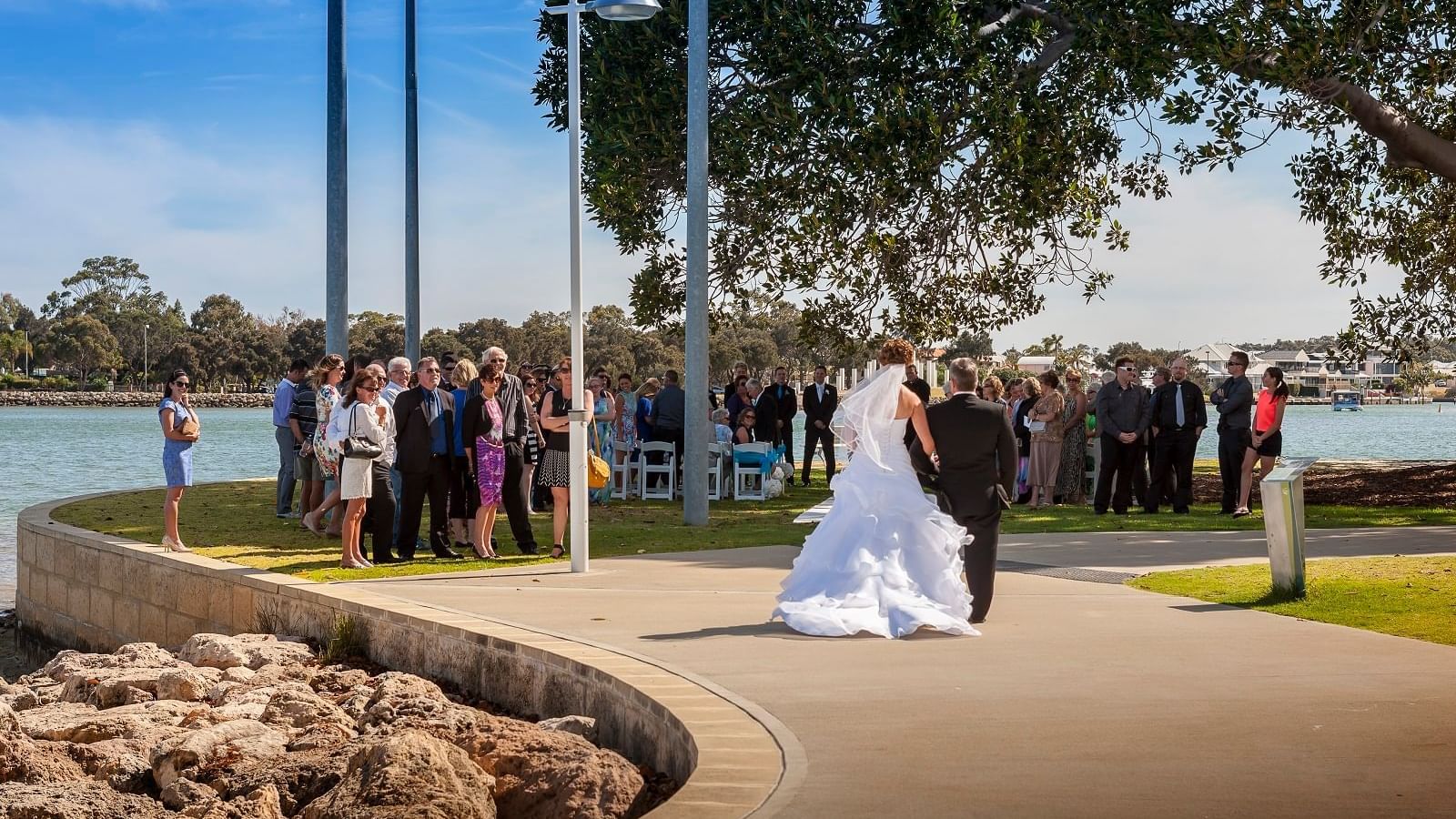 Bride and groom walk along an estuary path as wedding guests gather on a sunny day at The Sebel Mandurah