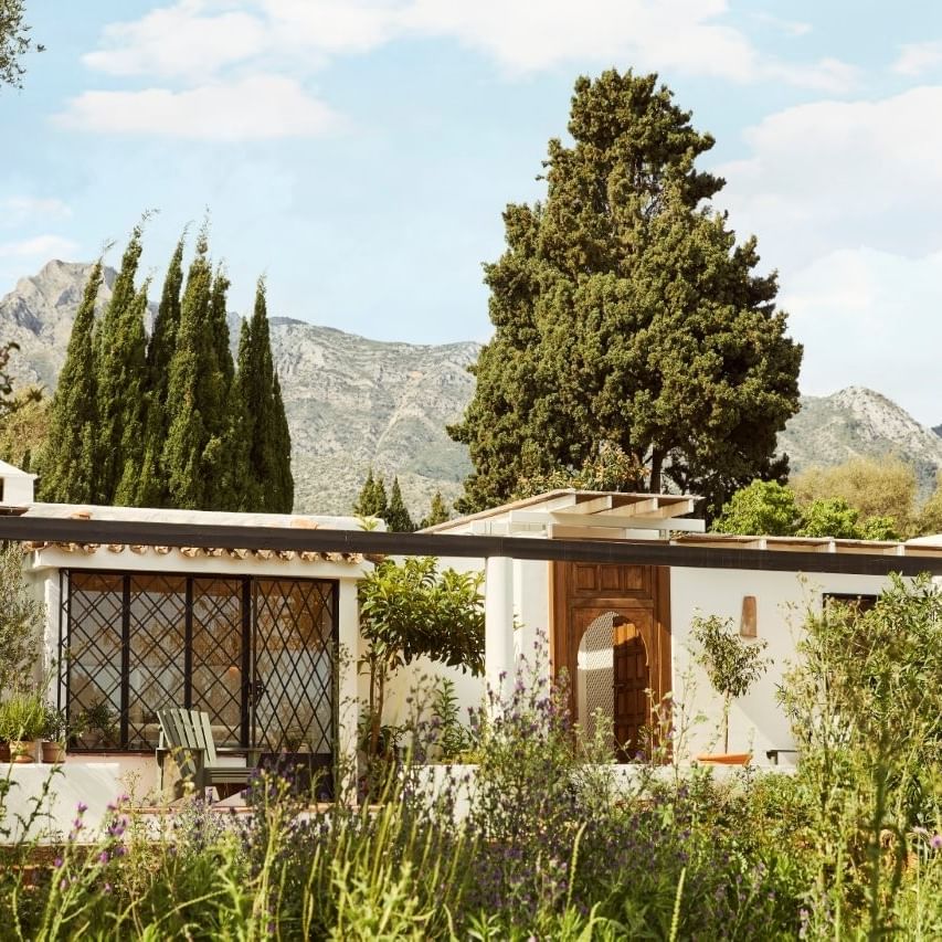 A white rustic house with a garden and mountain view in the background under a partly cloudy sky.