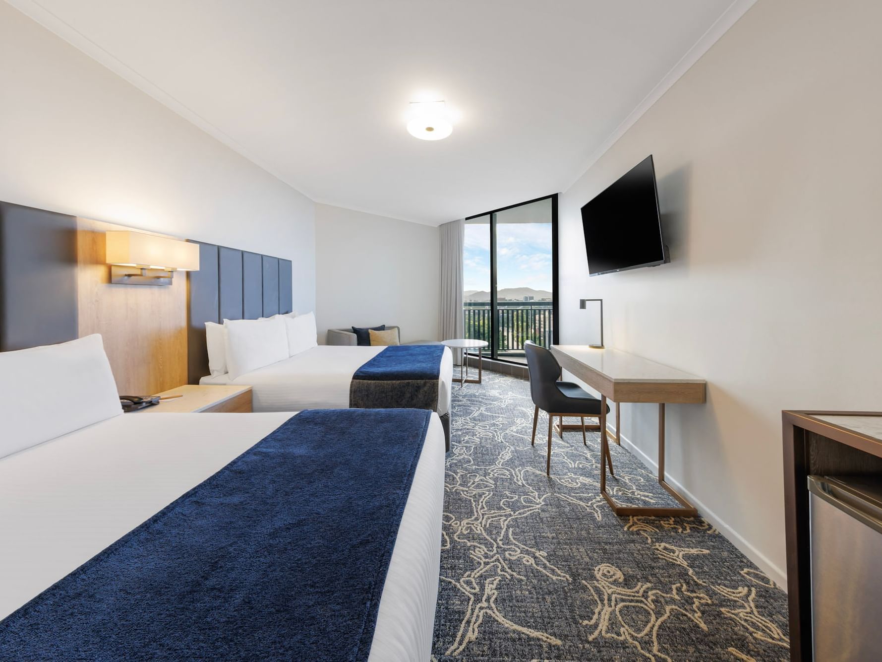 Modern twin room at Hotel Grand Chancellor Brisbane featuring two double beds with navy runners, a marble desk, and a large window with balcony access overlooking the city or Roma Street Parklands with Suncorp Stadium in the distance.