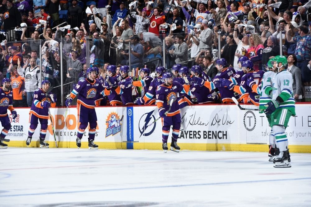 Hockey players in navy and orange and green and white uniforms on the ic in front of a large cheering crowd.