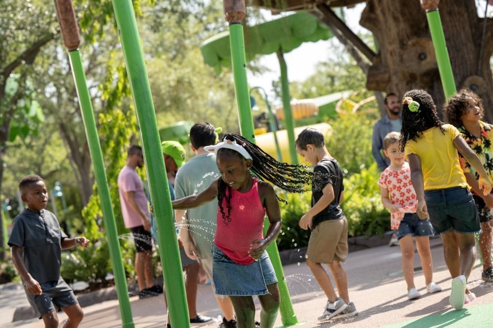 A girl in a tank top surrounded by other kids plays in a splash pad area with tall reed-like fountains.