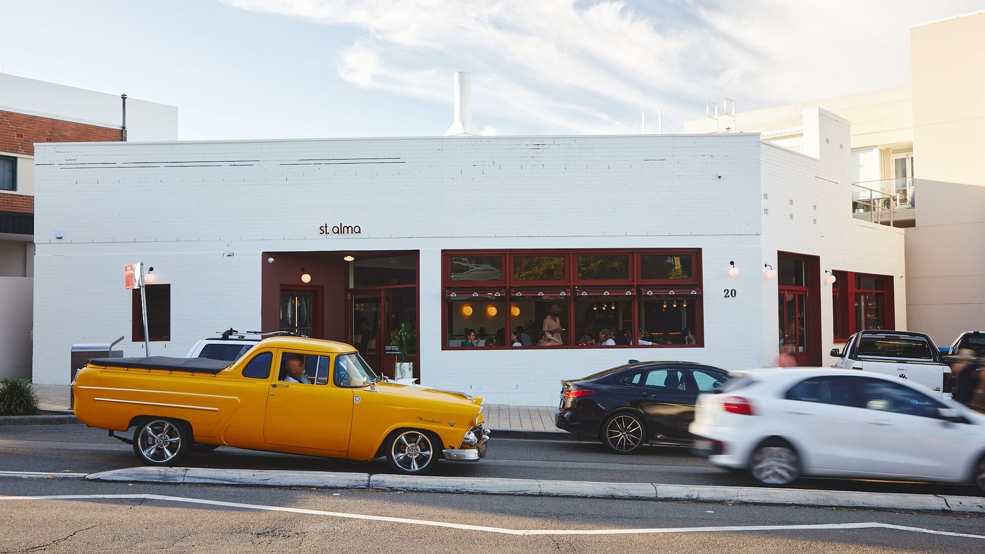 St. Alma with a Classic yellow truck driving by a white building under a bright blue sky near Elysium Manly