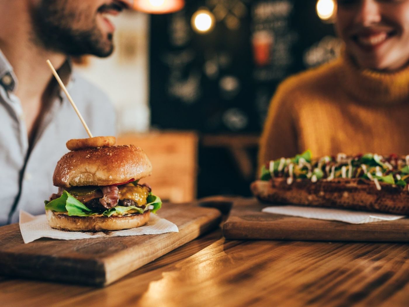 Hamburguesa de res sobre tabla de madera en The Grill de Fiesta Americana Funeeq Punta Cana, con pareja al fondo