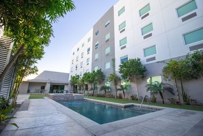 Clear pool surrounded by lounge chairs on a green lawn by a tall building under a bright sky at Hotel Mirra
