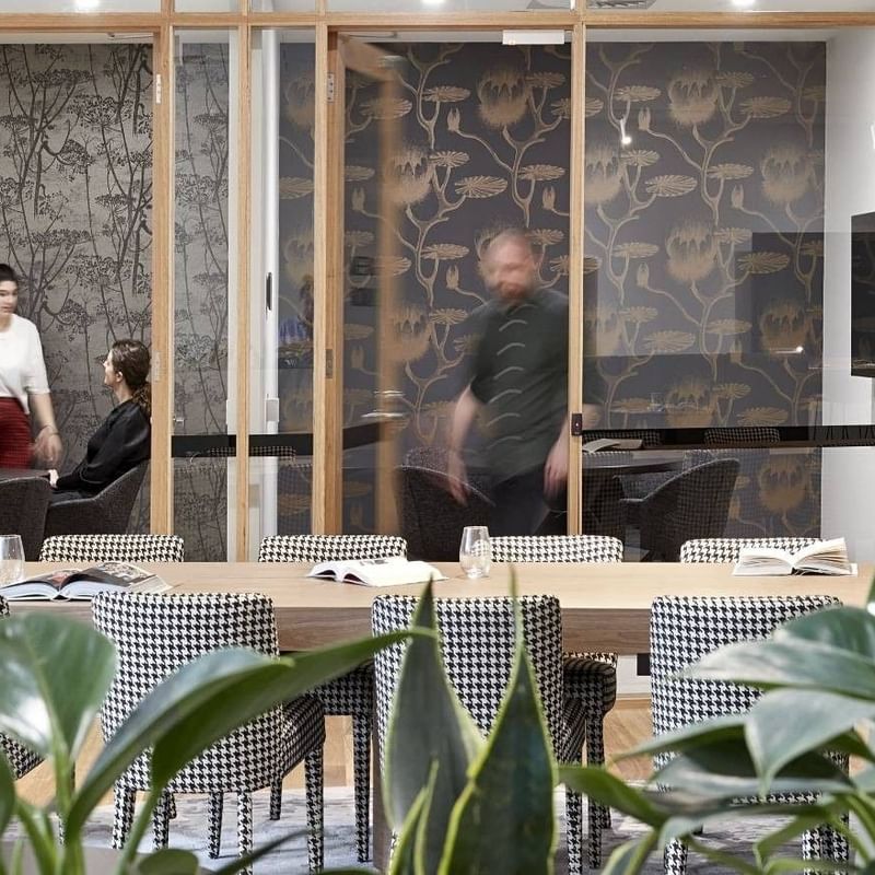 Office room with plants, a wooden table, and people near glass walls and a TV.
