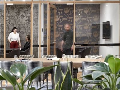 Office room with plants, a wooden table, and people near glass walls and a TV.