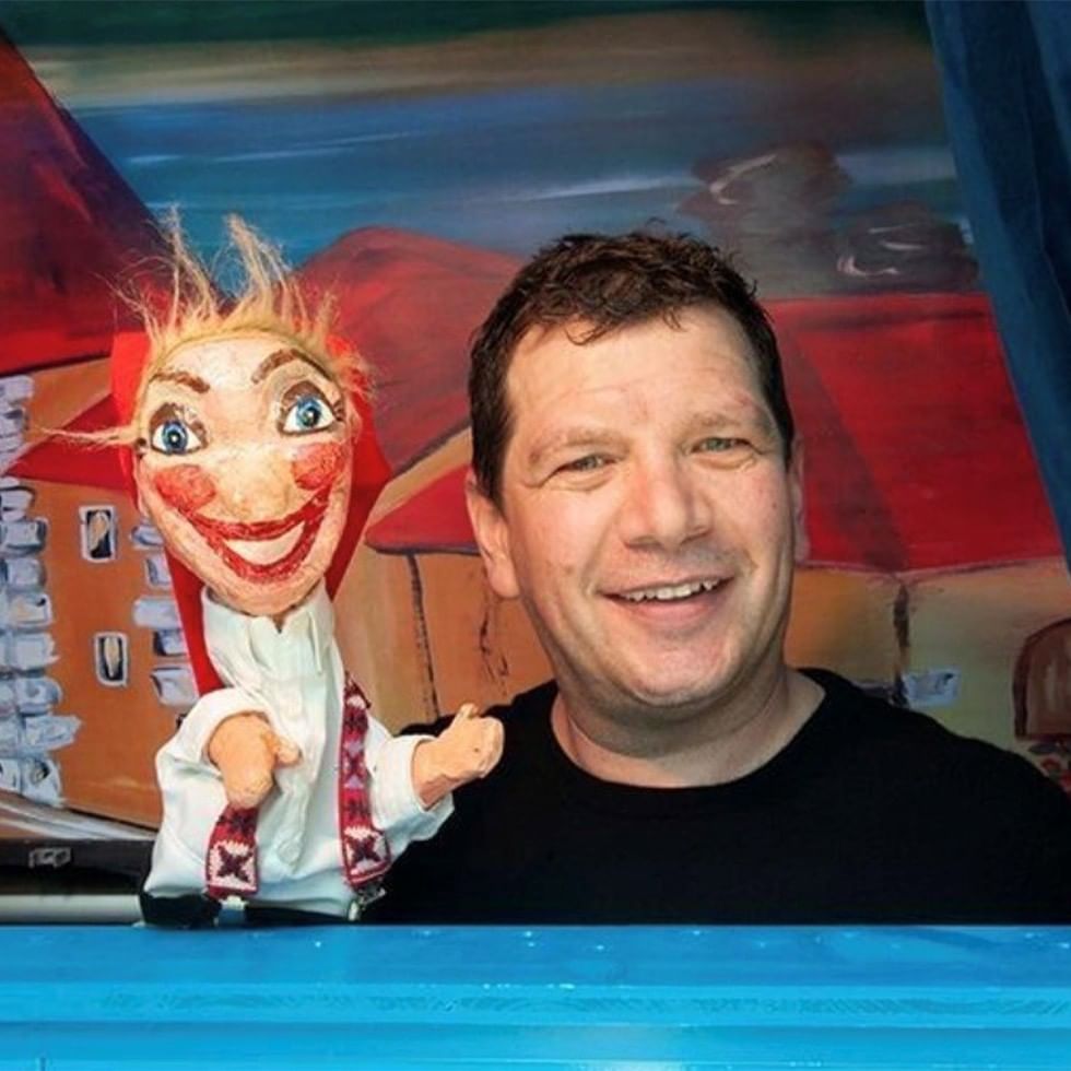 Lächelnder Mann mit Kasperlpuppe vor buntem Hintergrund beim Kasperltheater mit Christian Mair.