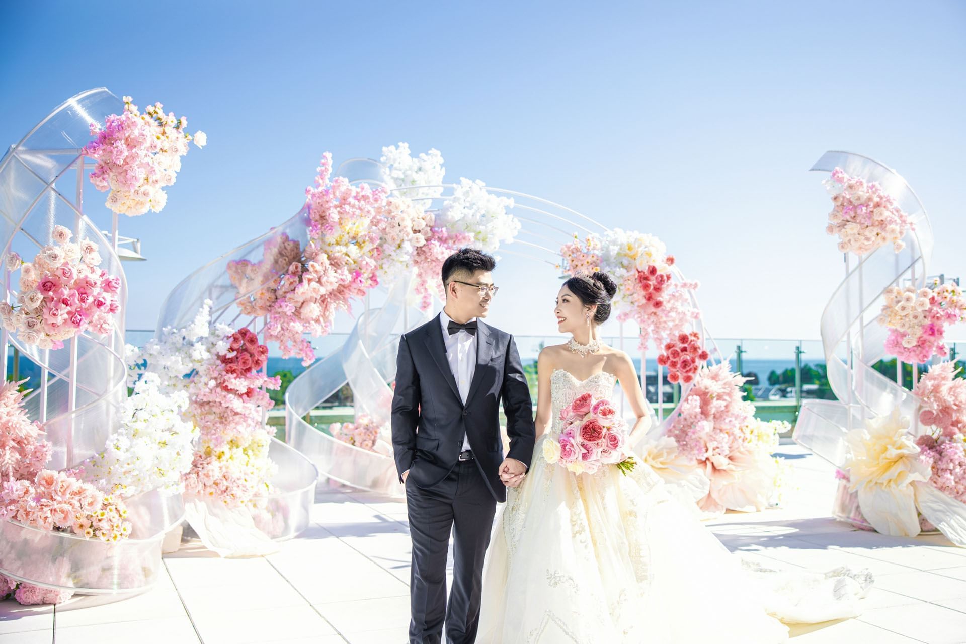 Bride and groom holding hands beside pink floral arches on an outdoor rooftop terrace facing forward at Hotel X Toronto