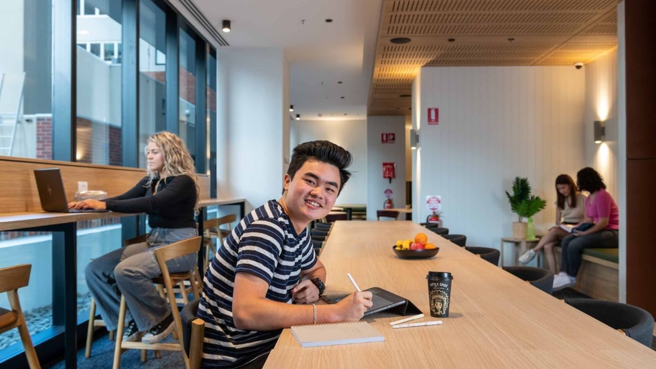 Smiling student holding a pen and a tablet, sitting at a table in study area with other students.