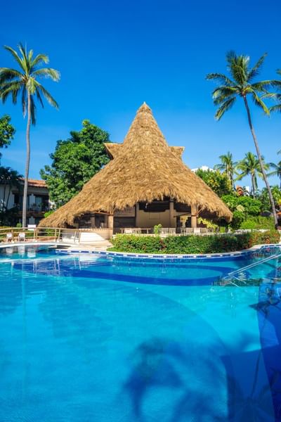 Resort pool with clear blue water, palm trees, and a thatched-roof hut under a bright sky.
