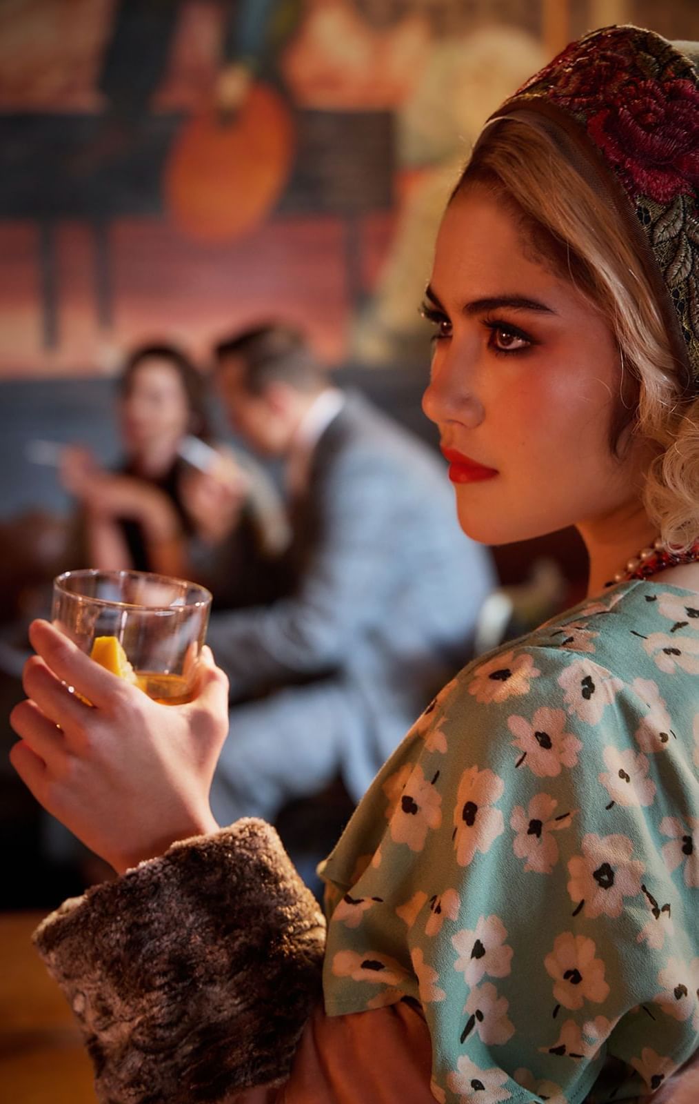 A woman with a headscarf and bright red lipstick holds a drink in a retro-themed The Midnight Bell at The Londoner Hotel