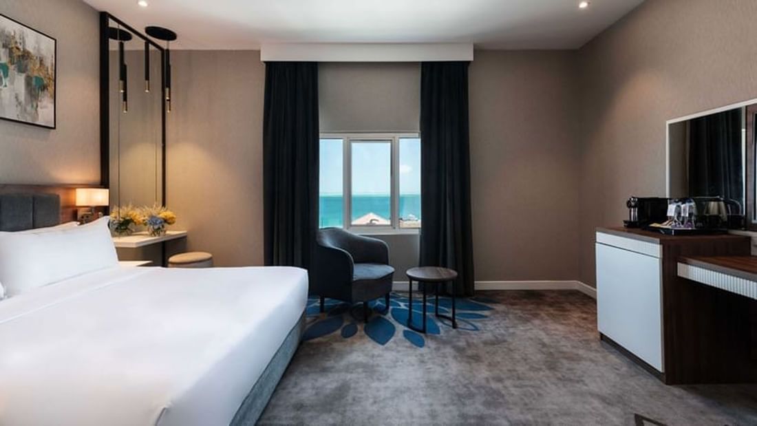 Dark armchair and table by a window near a white bed in Junior Suite Sea View at Warwick Al Jubail